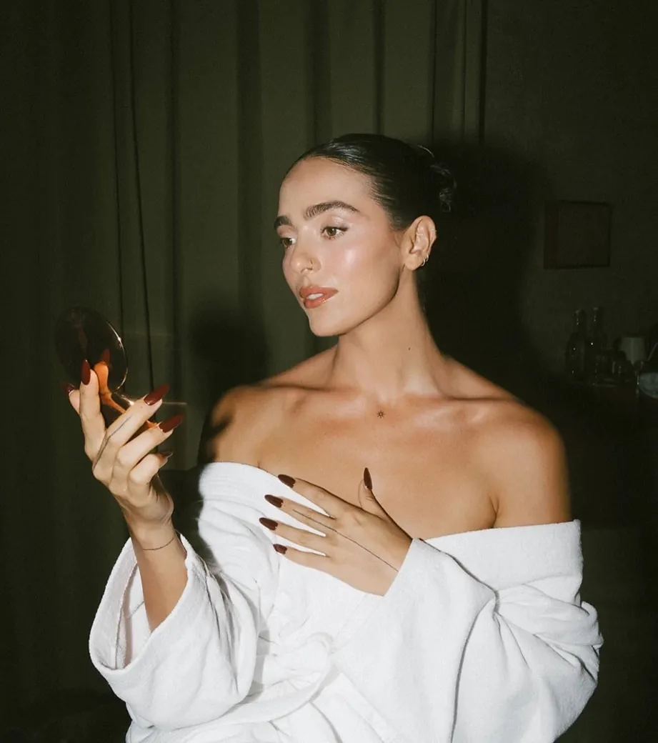 Model in a white robe checking her glowing skin in a hand mirror